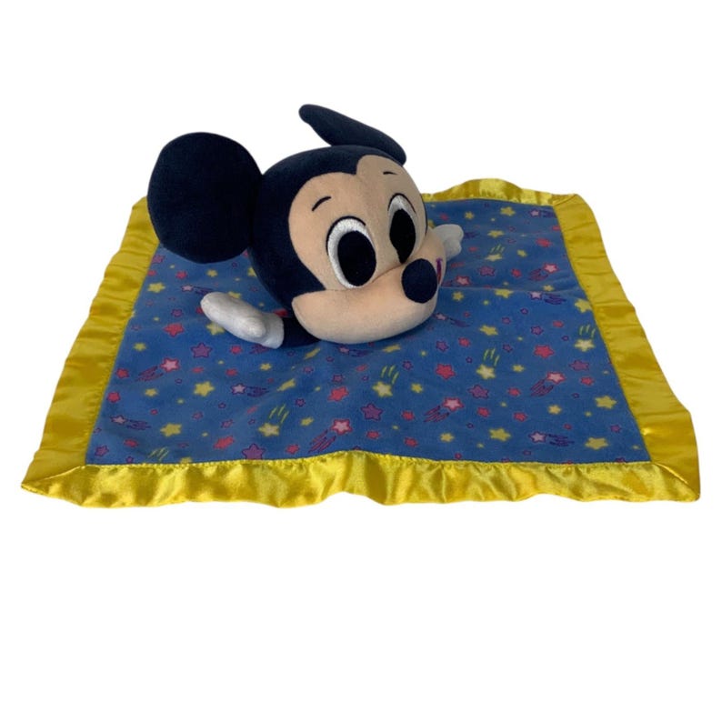 May include: A plush security blanket featuring a Mickey Mouse head and arms attached to a square blue fabric base. The blanket has a yellow satin border and a pattern of small yellow stars and pink flowers printed across the blue fabric surface.