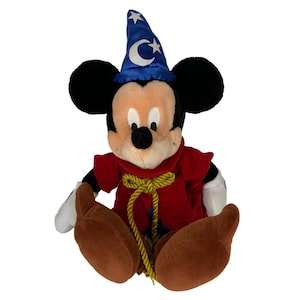 Peut inclure: Un Mickey Mouse en peluche portant un chapeau de magicien bleu avec des &eacute;toiles et une lune croissante. Il porte une robe rouge avec une corde dor&eacute;e attach&eacute;e autour de sa taille. Il est assis les jambes crois&eacute;es.