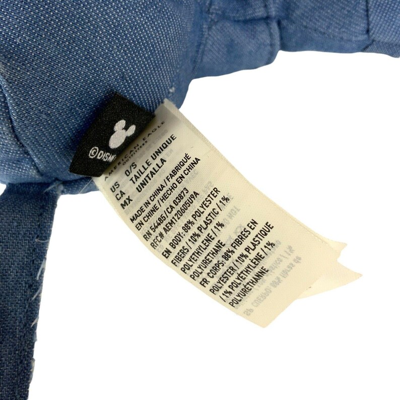 May include: A blue denim fabric item with a black tag featuring a white Mickey Mouse icon and a white care label. The label lists materials as 88% polyester, 10% plastic, 1% polyethylene, and 1% polyurethane, with manufacturing details from China.
