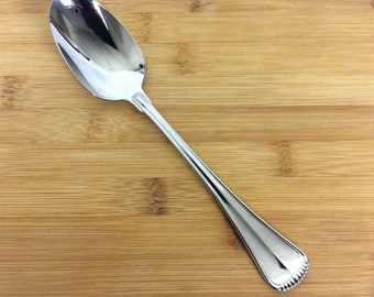 May include: A silver spoon with a rounded bowl and a long, slender handle. The spoon is lying on a wooden surface.