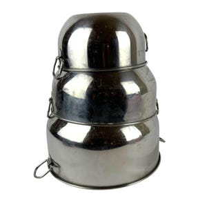 May include: A set of three stacked, polished stainless steel mixing bowls of varying sizes. Each bowl features a smooth, rounded exterior and a small, circular metal ring handle attached to the side for easy gripping and handling during use.