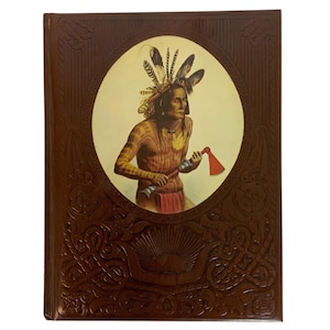 Puede incluir: Un libro vintage con una cubierta marrón con un diseño en relieve. El centro de la cubierta tiene una imagen circular de un hombre nativo americano sosteniendo un tomahawk. El hombre lleva un tocado de plumas y un taparrabos rojo.