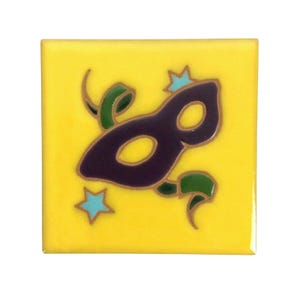 Op de afbeelding: Een gele keramische tegel met een ontwerp van een paars en gouden maskerade masker. Het masker is omgeven door groene en gouden swirls en blauwe sterren.