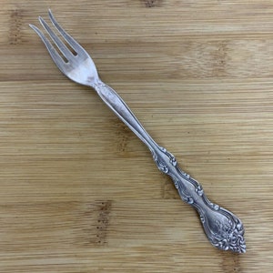 Può includere: Una forchetta d'argento con un manico decorato. La forchetta è su una superficie di legno.