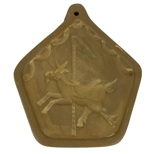 May include: A brown ceramic ornament shaped like a pentagon with a hole at the top for hanging. The ornament features a raised design of two horses on a carousel.