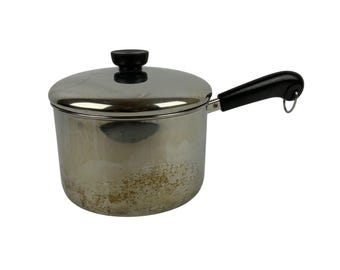 May include: A stainless steel saucepan with a polished finish and visible signs of wear on the lower exterior. It features a black plastic lid knob and a matching black handle with a small metal hanging loop at the end of the handle.