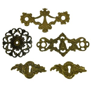 May include: Five ornate brass decorative hardware pieces with intricate designs. The pieces are in various shapes and sizes, including a circular piece with a central hole, a rectangular piece with a keyhole, and two smaller pieces with a keyhole.