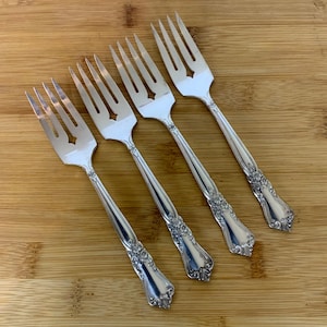 May include: Four silver forks with decorative floral patterns on the handles and a star-shaped cutout in the tines. The forks are displayed diagonally on a light brown wooden surface, highlighting their elegant design.