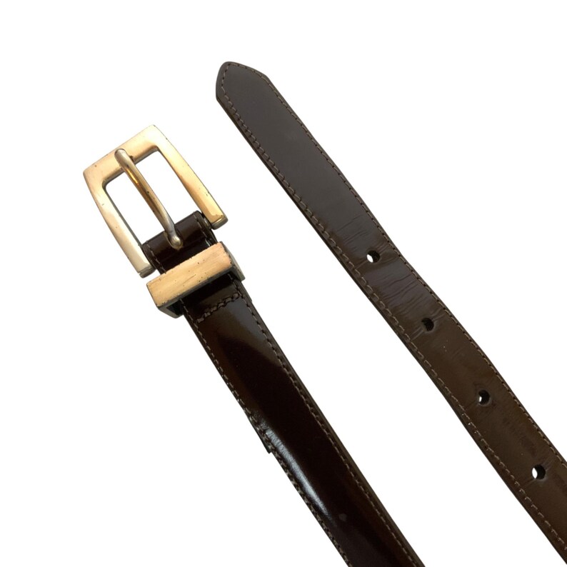 May include: A dark brown leather belt with a silver-colored buckle. The belt is glossy and has a rectangular buckle. The belt is unbuckled and the strap is separated from the buckle.