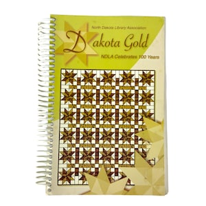 Può includere: Un quaderno a spirale con copertina gialla con la scritta "Dakota Gold" e "North Dakota Library Association". La copertina del quaderno presenta un motivo geometrico a stelle marroni e bianche, con la scritta "NDLA Celebrates 100 Years."