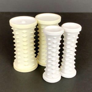 May include: Four white and cream-coloured cylindrical massage tools with textured surfaces and rounded tops. The tools vary in size, with the largest being approximately 15 cm tall. The textured surfaces feature evenly spaced, rounded protrusions.