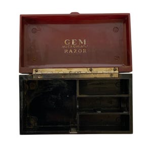 Puede incluir: Un estuche de afeitar vintage abierto, con tapa abisagrada. La tapa es de color marrón rojizo oscuro, y el interior es negro con compartimentos. El texto "GEM MICROMATIC RAZOR" está impreso en dorado en el interior de la tapa.