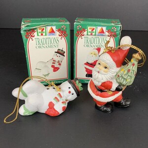 Op de afbeelding: Twee kerstversieringen: een sneeuwpop en de kerstman, met hun originele dozen. De sneeuwpop is wit met een rode sjaal en een zwarte hoed. De kerstman draagt een rood pak en houdt een groene kerstboom vast. De dozen zijn groen met de woorden "Traditions Ornament".