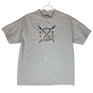 Könnte beinhalten: Graues, gestreiftes T-Shirt mit einem gestickten Logo, das gekreuzte Ruder, einen Anker und den Text "St. Augustine FL USA" zeigt.