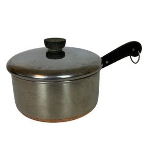 May include: A stainless steel saucepan with a lid and a black handle. The pot has a copper-coloured base and a black knob on the lid. The handle has a hole for hanging.