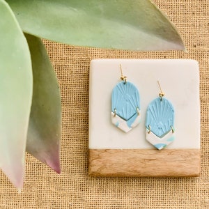 May include: A pair of light blue and white earrings with gold-colored hooks. The earrings have a geometric design with a textured, fan-like pattern on the top portion. The earrings are displayed on a white and wood-toned rectangular surface.