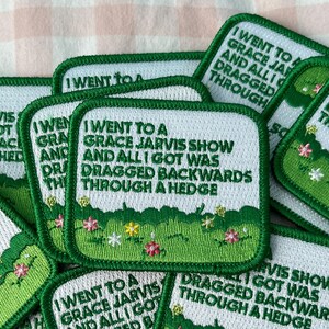 May include: A collection of square embroidered patches featuring a white background with green text that reads I WENT TO A GRACE JARVIS SHOW AND ALL I GOT WAS DRAGGED BACKWARDS THROUGH A HEDGE, with a green hedge and small colorful flowers at the bottom.