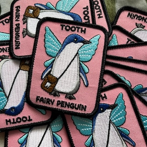 May include: A rectangular embroidered patch with a pink background featuring a penguin with light blue fairy wings and a brown satchel. The text TOOTH FAIRY PENGUIN is stitched in black lettering. The patch has a thick black border.