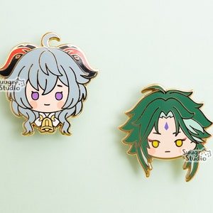 May include: Two enamel pins featuring anime characters. The first pin is a light blue character with red and black horns, a white collar, and a gold bell. The second pin is a green character with a purple gem on their forehead. Both pins have gold outlines and are on a light green background. The text "Swag Studio" is visible on both pins.