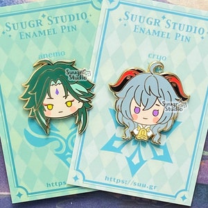 May include: Two enamel pins featuring anime characters. The pins are on cards with the text "SUUGR STUDIO. ENAMEL PIN" and the character's element. The left pin features a green-haired character with a purple gem on their forehead. The right pin features a blue-haired character with a red horn and a bell around their neck.