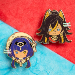 Può includere: Due spille in smalto dorate con personaggi in stile anime. La prima spilla raffigura un personaggio con lunghi capelli neri, occhi gialli e un abito rosso e oro. La seconda spilla raffigura un personaggio con capelli blu, occhi gialli e un abito blu e oro. Entrambe le spille hanno uno sfondo bianco con il testo "sugar studio" in rosa e blu.