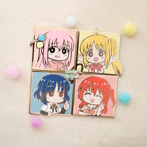 May include: Four square enamel pins featuring cartoon characters with different coloured hair and clothing. The characters are smiling and have large eyes. The pins are gold-coloured and have a glossy finish.