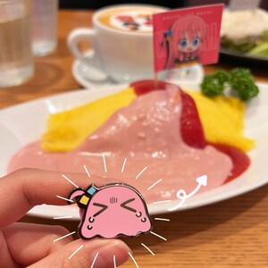 Puede incluir: Un pin de esmalte rosa y amarillo con un personaje de dibujos animados con una expresi&oacute;n triste. El personaje tiene una cabeza grande, ojos peque&ntilde;os y una boca con un ce&ntilde;o fruncido. El pin est&aacute; siendo sostenido por una mano frente a un plato de comida.