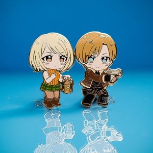 K&ouml;nnte beinhalten: Zwei goldfarbene Emaille-Pins mit zwei Anime-Charakteren. Die weibliche Figur hat blonde Haare, einen gr&uuml;nen karierten Rock und h&auml;lt eine Laterne. Die m&auml;nnliche Figur hat braunes Haar, eine braune Jacke und h&auml;lt eine Pistole. Beide Figuren haben gro&szlig;e Augen und l&auml;cheln.