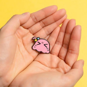 Puede incluir: Pin de esmalte rosa con un personaje de dibujos animados con una expresi&oacute;n triste. El personaje tiene una diadema azul y amarilla y tiene forma de mancha.