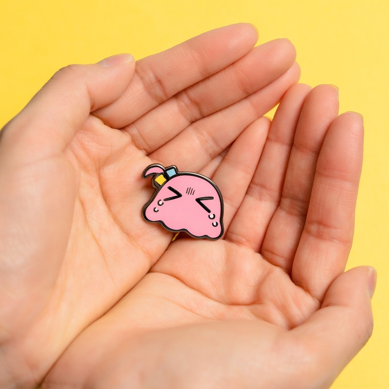 Puede incluir: Pin de esmalte rosa con un personaje de dibujos animados con una expresi&oacute;n triste. El personaje tiene una diadema azul y amarilla y tiene forma de mancha.
