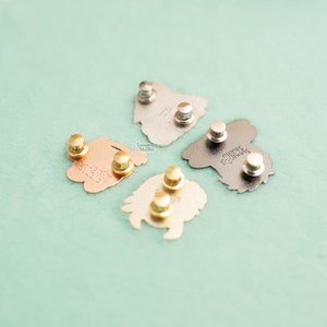 May include: A collection of enamel pins in various metallic finishes, including gold, silver, and copper. The backs of the pins are visible, showing the pin backs and the "Suugree Studio" logo. The pins are arranged on a light green surface.