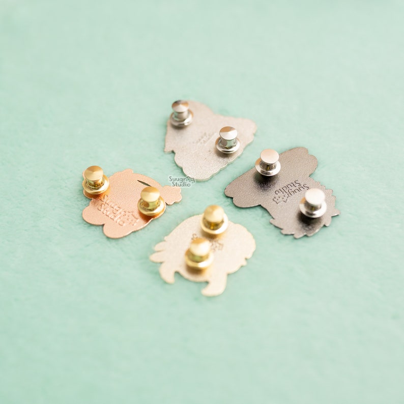 May include: A collection of enamel pins in various metallic finishes, including gold, silver, and copper. The backs of the pins are visible, showing the pin backs and the "Suugree Studio" logo. The pins are arranged on a light green surface.