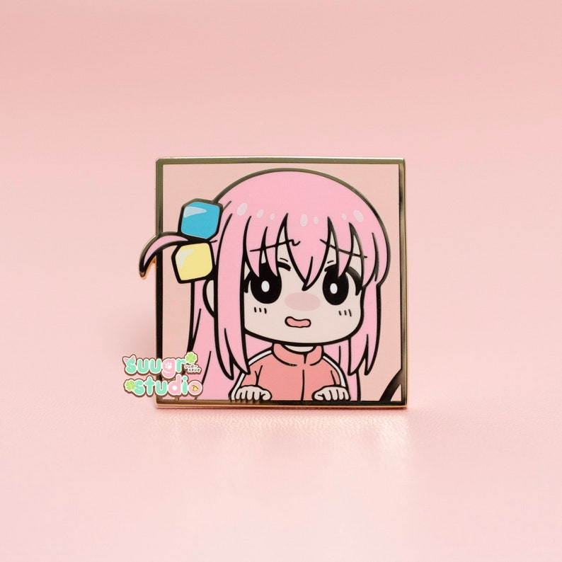 May include: A pink enamel pin featuring a cartoon character with pink hair, a pink shirt, and a surprised expression. The character is wearing a blue and yellow bow. The pin is set against a gold background.