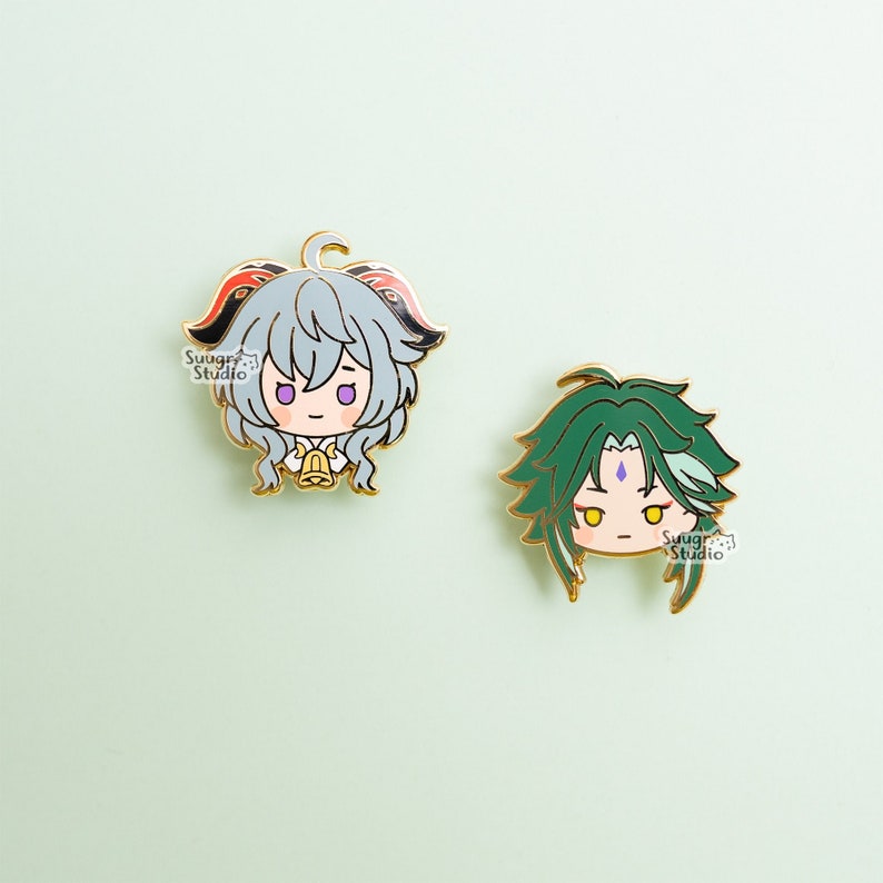 May include: Two enamel pins featuring anime characters. The first pin is a light blue character with red and black horns, a white collar, and a gold bell. The second pin is a green character with a purple gem on their forehead. Both pins have gold outlines and are on a light green background. The text "Swag Studio" is visible on both pins.