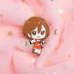 Puede incluir: Un pin de esmalte dorado con un personaje de dibujos animados con cabello casta&ntilde;o y un atuendo rojo. El personaje sonr&iacute;e y parece estar en movimiento. El pin est&aacute; sobre un fondo de tela rosa con adornos de estrellas.