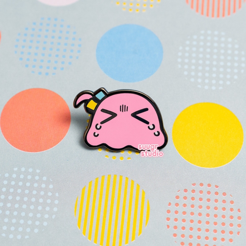 Puede incluir: Pin de esmalte rosa de un personaje de blob de dibujos animados con una expresi&oacute;n triste. El blob tiene un peque&ntilde;o lazo azul y amarillo en la cabeza. El pin dice "sugar studio" en la parte inferior.