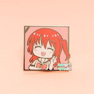 May include: A gold-coloured enamel pin featuring a cartoon character with red hair, wearing a yellow and white uniform with a red bow. The character is smiling and giving a peace sign. The pin is set against a pink background and has the text "sugar studio" in green and white.