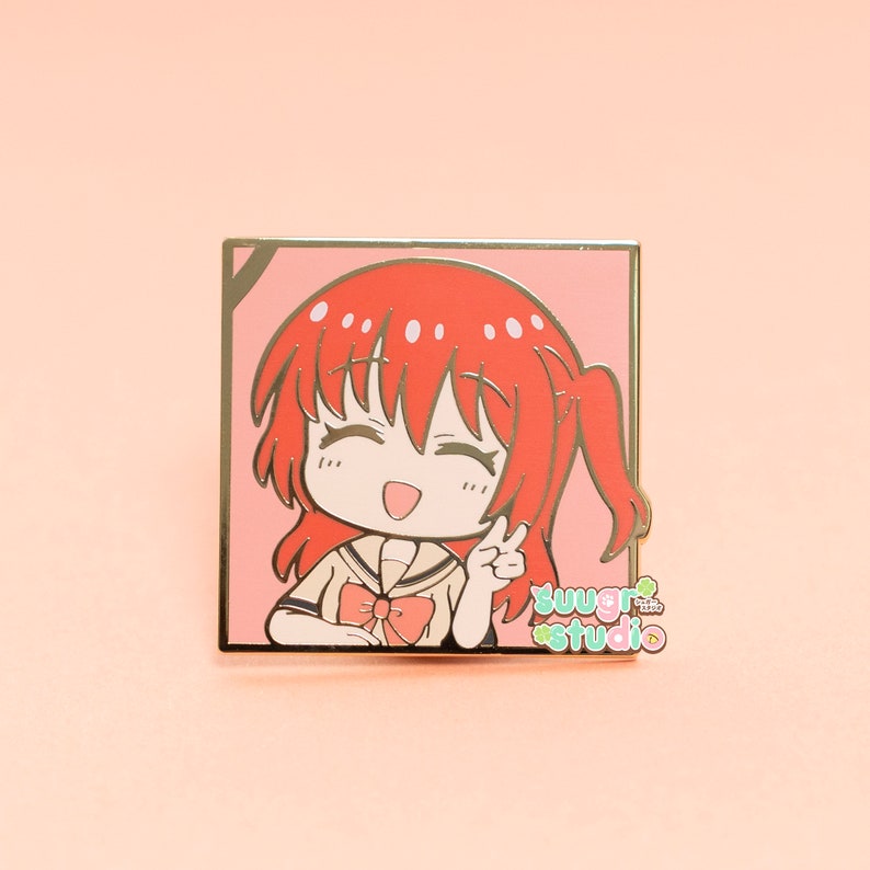 May include: A gold-coloured enamel pin featuring a cartoon character with red hair, wearing a yellow and white uniform with a red bow. The character is smiling and giving a peace sign. The pin is set against a pink background and has the text "sugar studio" in green and white.
