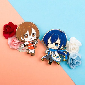 Puede incluir: Dos pines de esmalte con dise&ntilde;os de personajes de estilo anime. Uno muestra un personaje con cabello casta&ntilde;o y un atuendo naranja, el otro tiene cabello azul y un atuendo blanco y azul. Los pines se muestran con flores en el fondo.