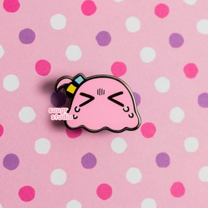 Puede incluir: Pin de esmalte rosa con una cara de dibujos animados con una expresi&oacute;n triste. La cara tiene un peque&ntilde;o lazo azul y amarillo en la parte superior. El pin est&aacute; sobre un fondo rosa con lunares blancos y morados.