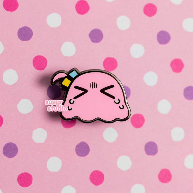 Puede incluir: Pin de esmalte rosa con una cara de dibujos animados con una expresi&oacute;n triste. La cara tiene un peque&ntilde;o lazo azul y amarillo en la parte superior. El pin est&aacute; sobre un fondo rosa con lunares blancos y morados.