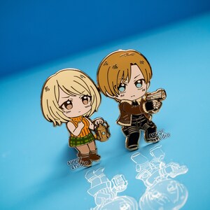 K&ouml;nnte beinhalten: Goldfarbene Emaille-Pins mit einem Chibi-Anime-Paar. Die weibliche Figur hat blonde Haare und h&auml;lt eine Laterne. Die m&auml;nnliche Figur hat braunes Haar und h&auml;lt eine Pistole. Beide Figuren tragen Freizeitkleidung.