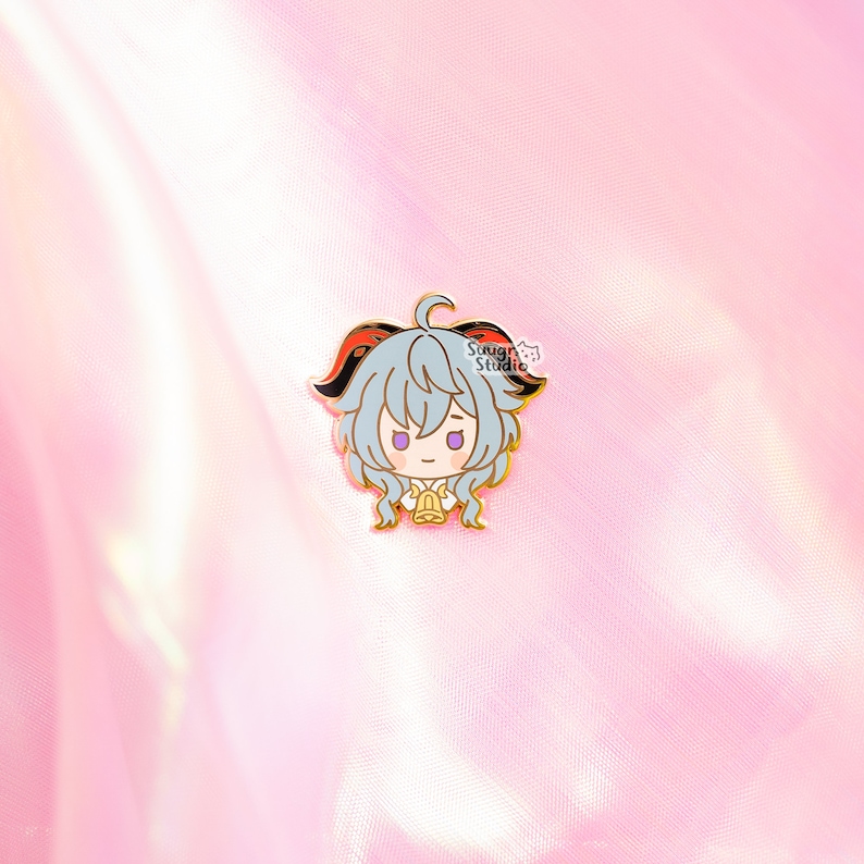 May include: A gold-colored enamel pin featuring a cartoon character with gray hair, red horns, and a white shirt. The character is holding a bell in their hands. The pin has a white background with the text "Sough Studio" in black.