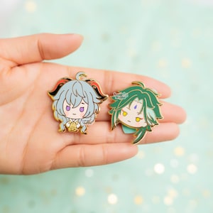 May include: Two gold-colored enamel pins featuring anime-style characters. The pin on the left features a character with light blue hair, red horns, and a bell. The pin on the right features a character with green hair and a purple gem on their forehead. Both pins have the text "Sugar Studio" on them.