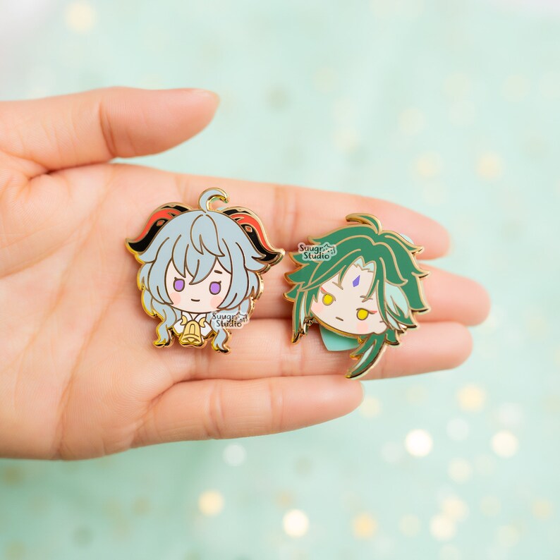 May include: Two gold-colored enamel pins featuring anime-style characters. The pin on the left features a character with light blue hair, red horns, and a bell. The pin on the right features a character with green hair and a purple gem on their forehead. Both pins have the text "Sugar Studio" on them.