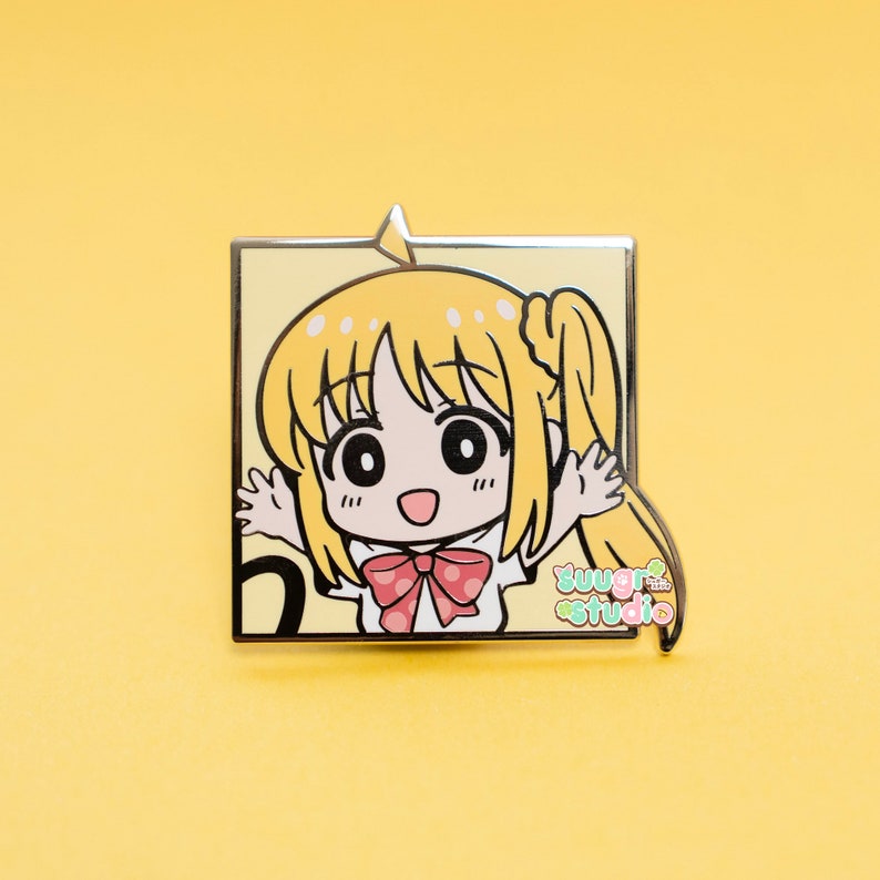 May include: A yellow and white enamel pin featuring a cartoon character with blonde hair, a red and white polka dot bow, and the text "sugar studio".