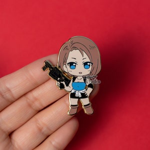May include: A gold-colored enamel pin featuring a chibi-style character with brown hair and blue eyes, holding a black gun. The character wears a blue top, black shorts, and brown boots. The pin has the text "suugr studio".