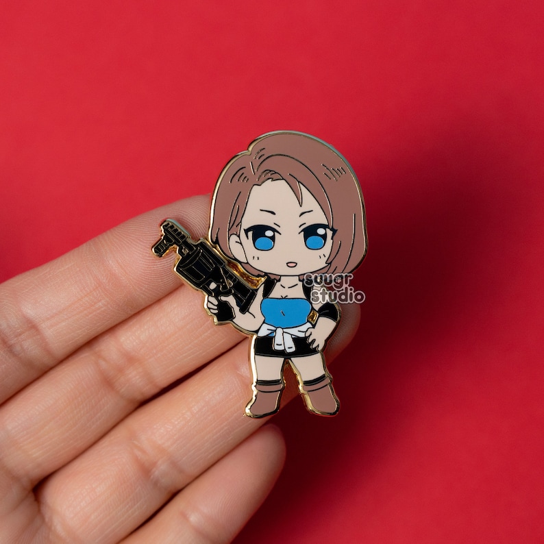 May include: A gold-colored enamel pin featuring a chibi-style character with brown hair and blue eyes, holding a black gun. The character wears a blue top, black shorts, and brown boots. The pin has the text "suugr studio".