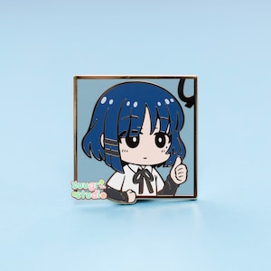 May include: A blue and white enamel pin featuring a cartoon character with blue hair, a white shirt, and a black bow tie. The character is giving a thumbs up. The pin has a gold border and the text "sugar studio" is printed in the bottom left corner.