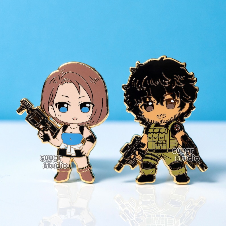 May include: Two enamel pins featuring chibi-style characters. One pin depicts a woman with brown hair and a blue top holding a gun. The other pin shows a man in military attire, also holding a gun. Both pins have gold-colored outlines and the text "suugr studio".